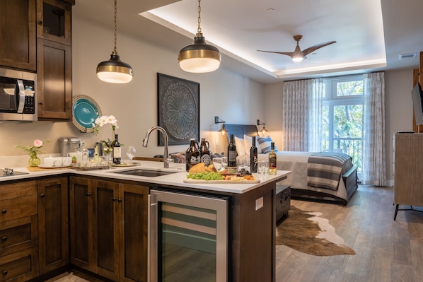 A modern kitchen with a central island, a dining area with a buffet setup, a bedroom with a bed and curtains, and a ceiling fan.