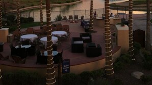 A patio area with round tables and chairs, surrounded by palm trees, overlooking a body of water and mountains.