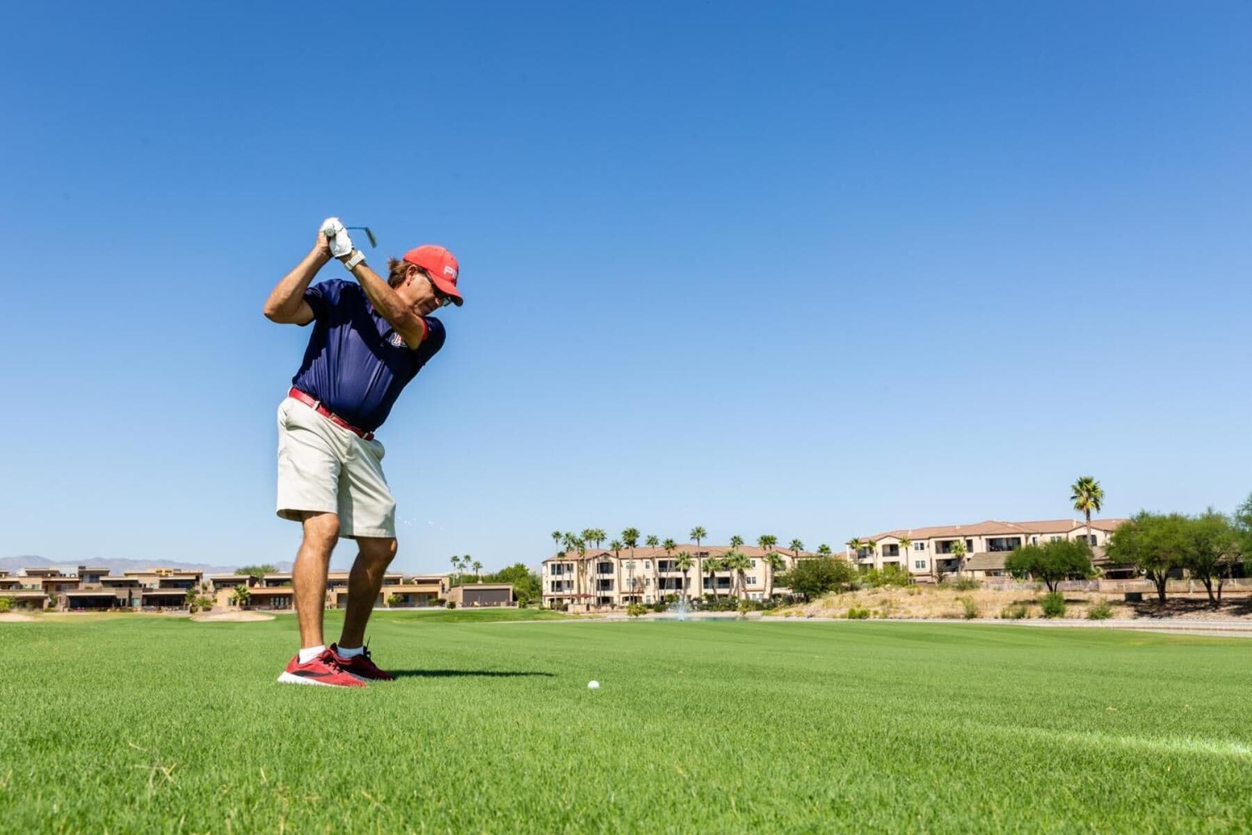 A golf course with a clear sky, a golfer in mid-swing, and a building complex in the background.