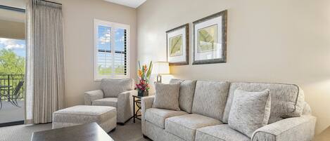 A living room with a sofa, armchair, and a coffee table. There is a window with blinds, a lamp, and framed pictures on the wall.