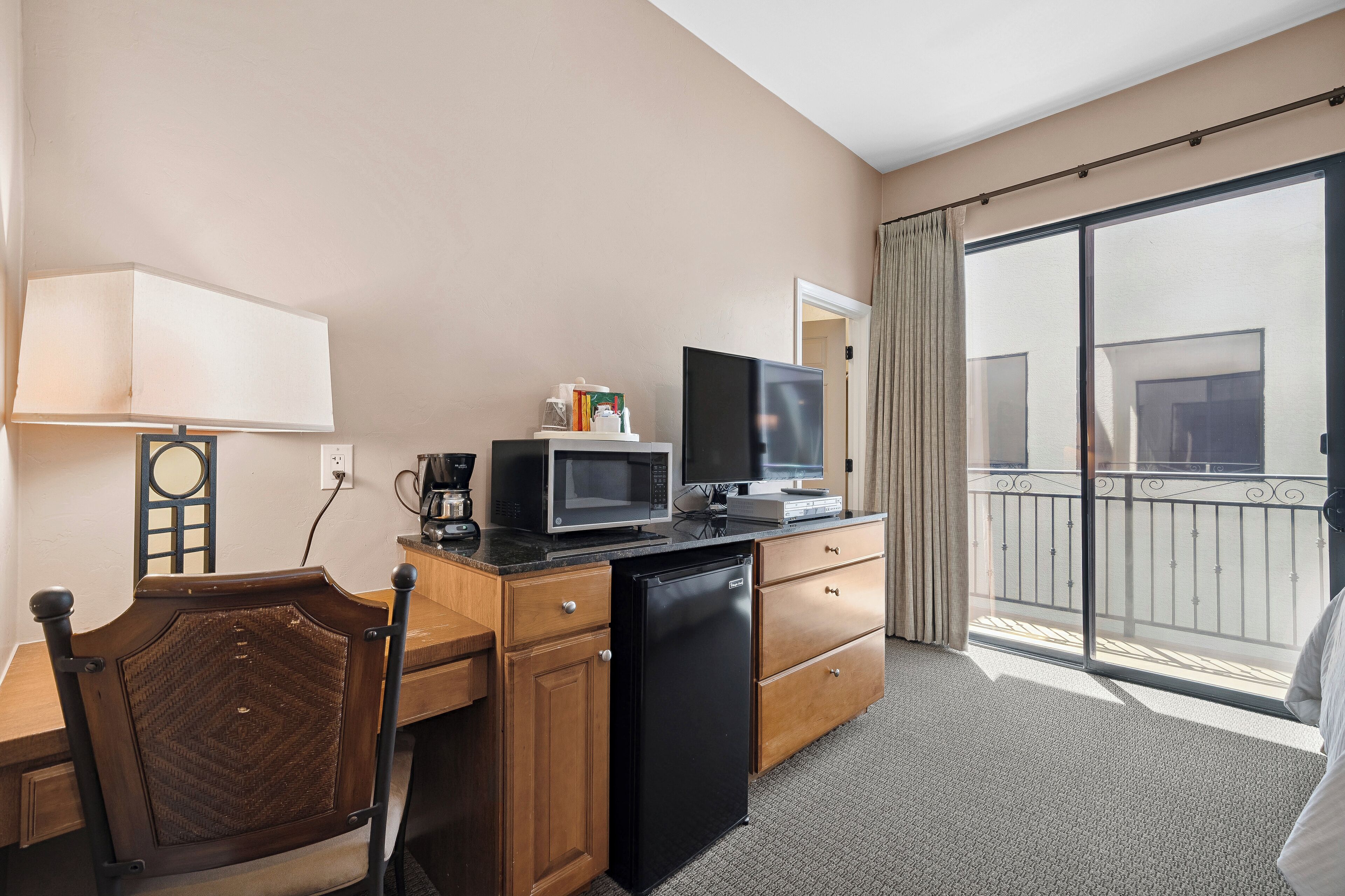 Habitación de hotel con kitchenette, un televisor de pantalla plana, refrigerador, microondas, cafetera, una silla de madera y un amplio ventanal corredero al balcón.