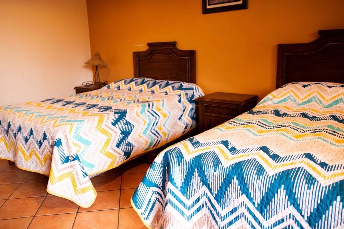 Una habitación con dos camas, cada una con una colcha estampada, cabeceros de madera y una mesita de noche con lámpara.