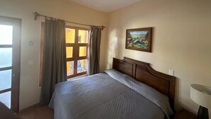 Un dormitorio con cama, ventilador de techo, ventana con cortinas, un cuadro en la pared y una lámpara en la mesita de noche.