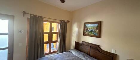 A bedroom with a bed, a ceiling fan, a window with curtains, a painting on the wall, and a lamp on a bedside table.
