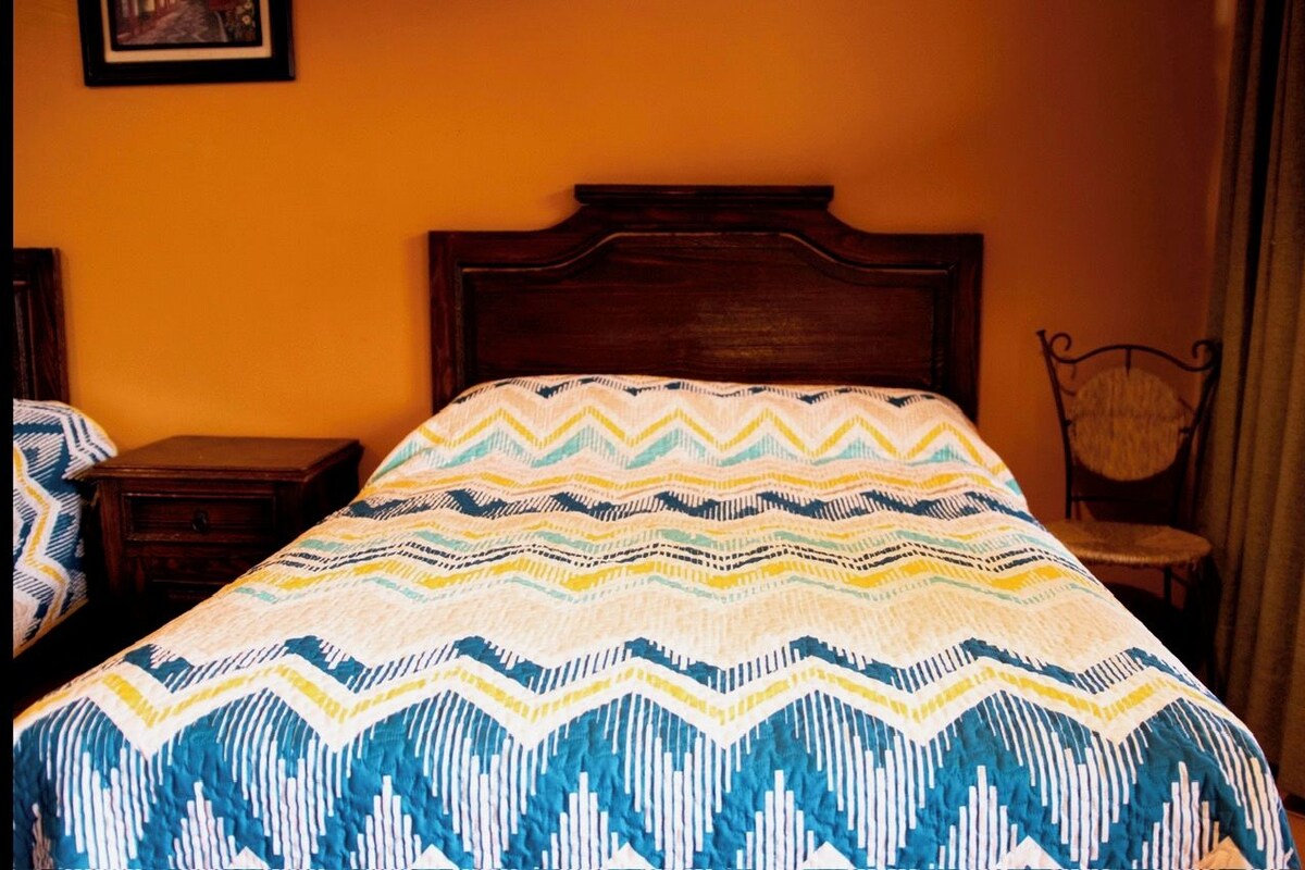 Una cama con una colcha estampada, un cabecero de madera, una mesita de noche y una silla de mimbre.
