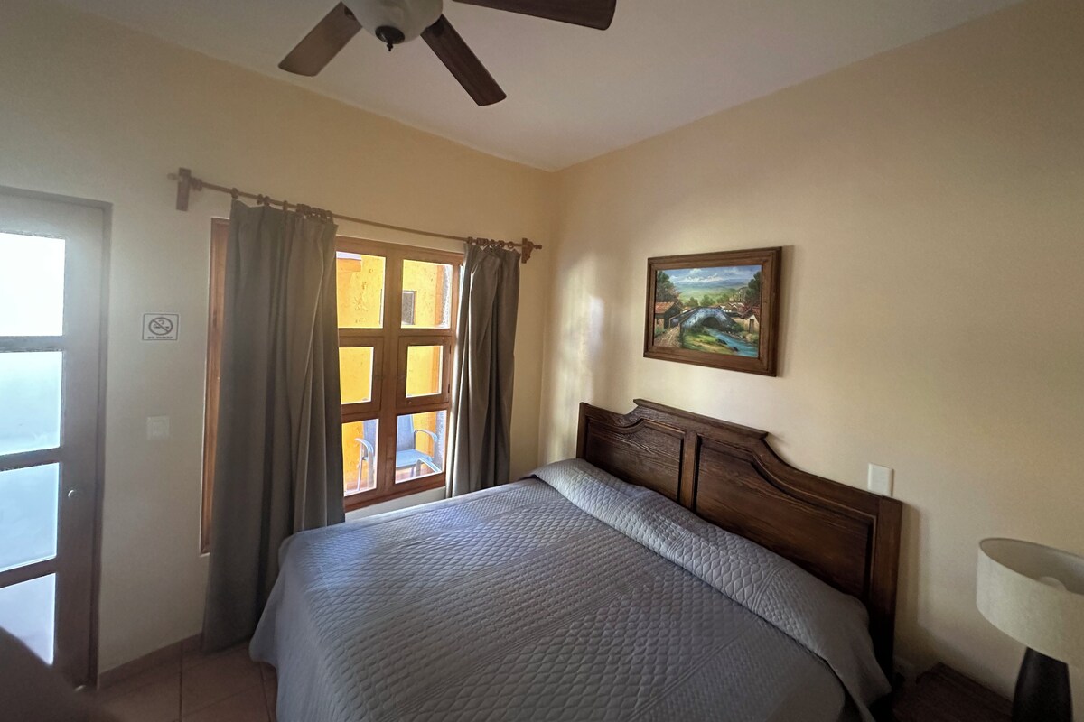 Un dormitorio con cama, ventilador de techo, ventana con cortinas y un cuadro en la pared.