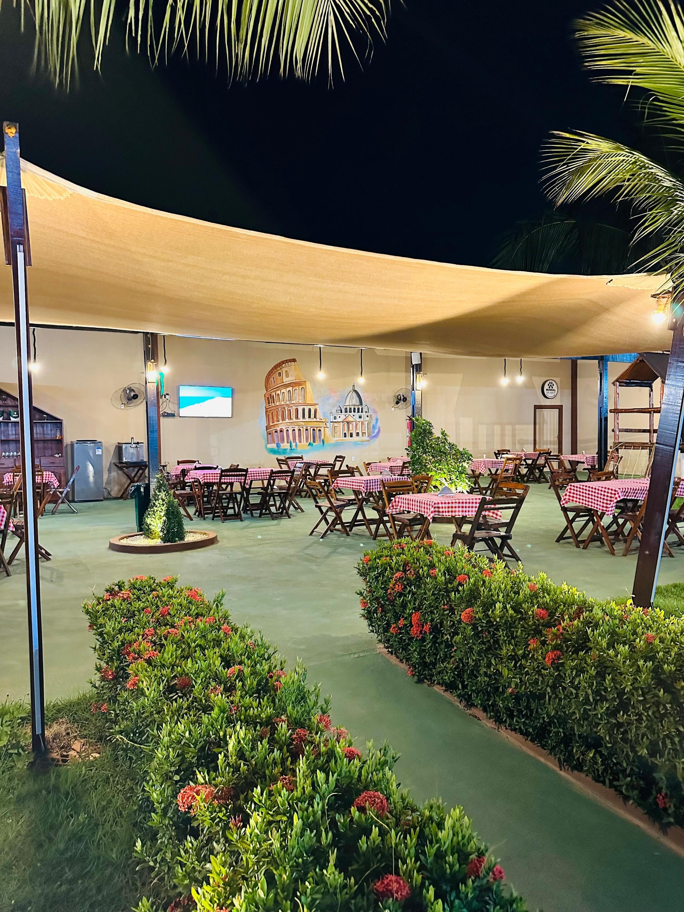 An outdoor dining area with tables covered in red and white checkered tablecloths, surrounded by greenery and a large mural of famous landmarks.