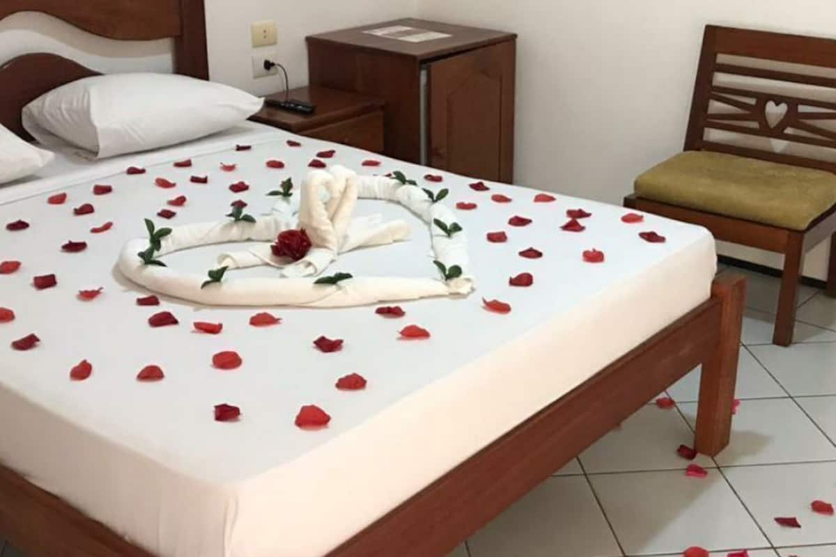 A neatly made bed with rose petals and a towel arrangement, a wooden chair, and a small wooden nightstand with a lamp.