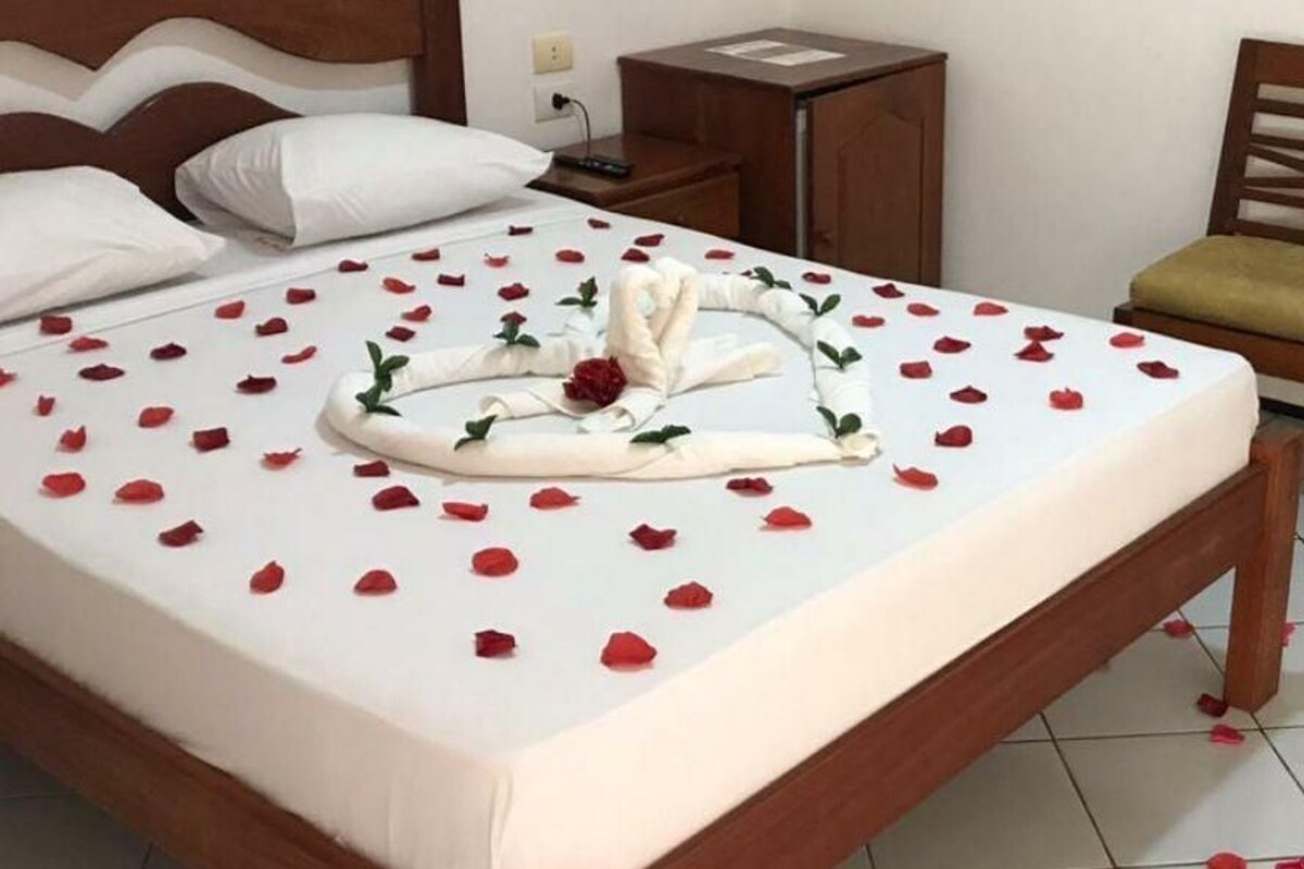 A neatly made bed with rose petals and a towel arrangement, a wooden chair, a nightstand, and a small cabinet.