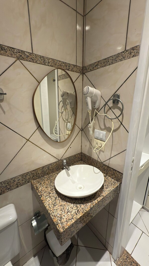 A bathroom with a granite countertop sink, a round mirror, and a hairdryer mounted on the wall.