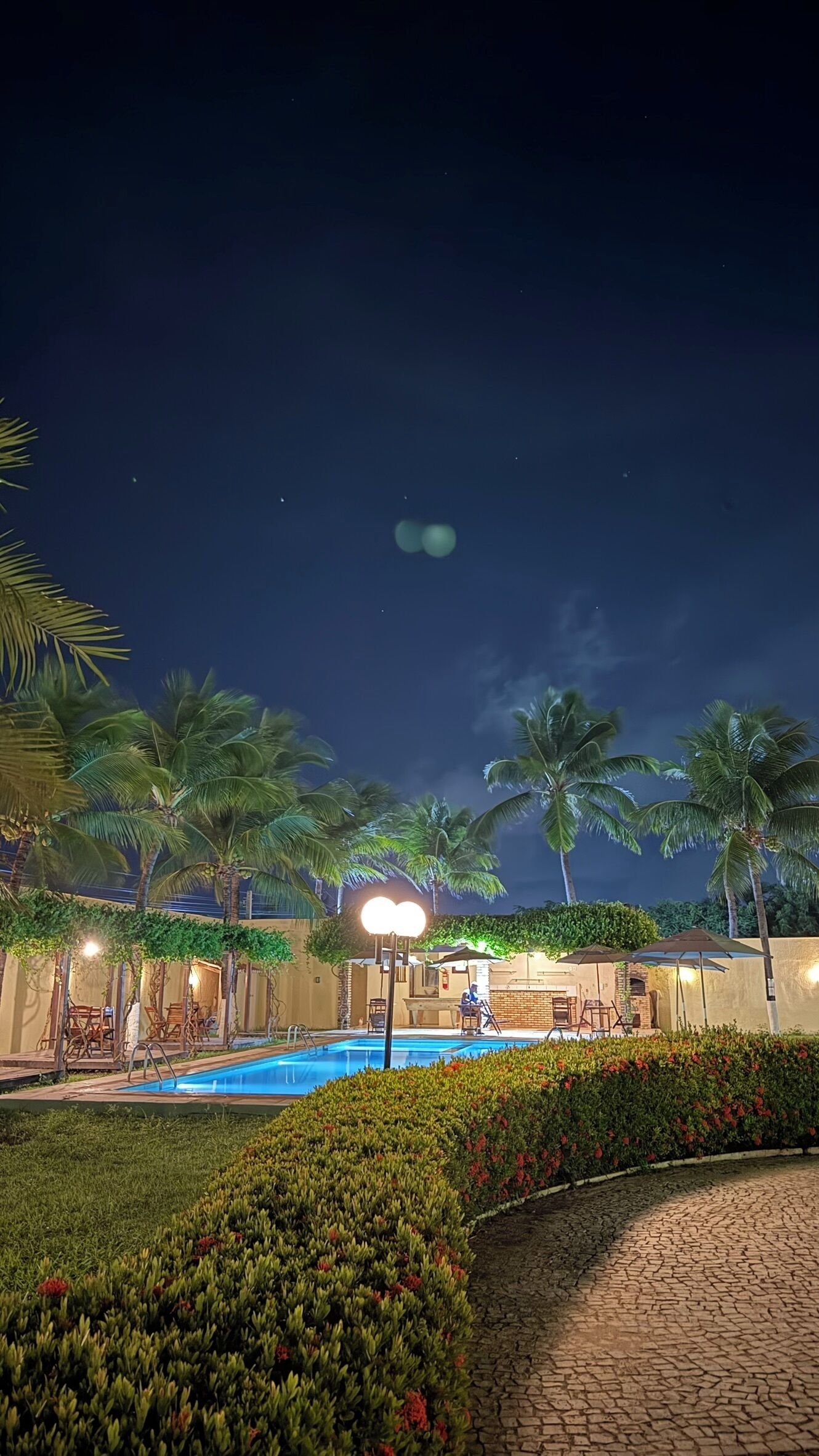 Vista nocturna de un resort con piscina, palmeras y zonas de estar al aire libre.