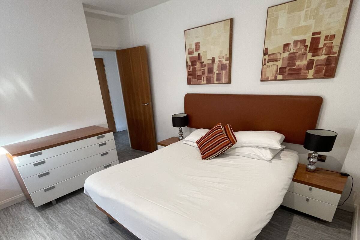 A modern bedroom with a large bed, wooden headboard, two bedside tables with lamps, a dresser, and abstract wall art.
