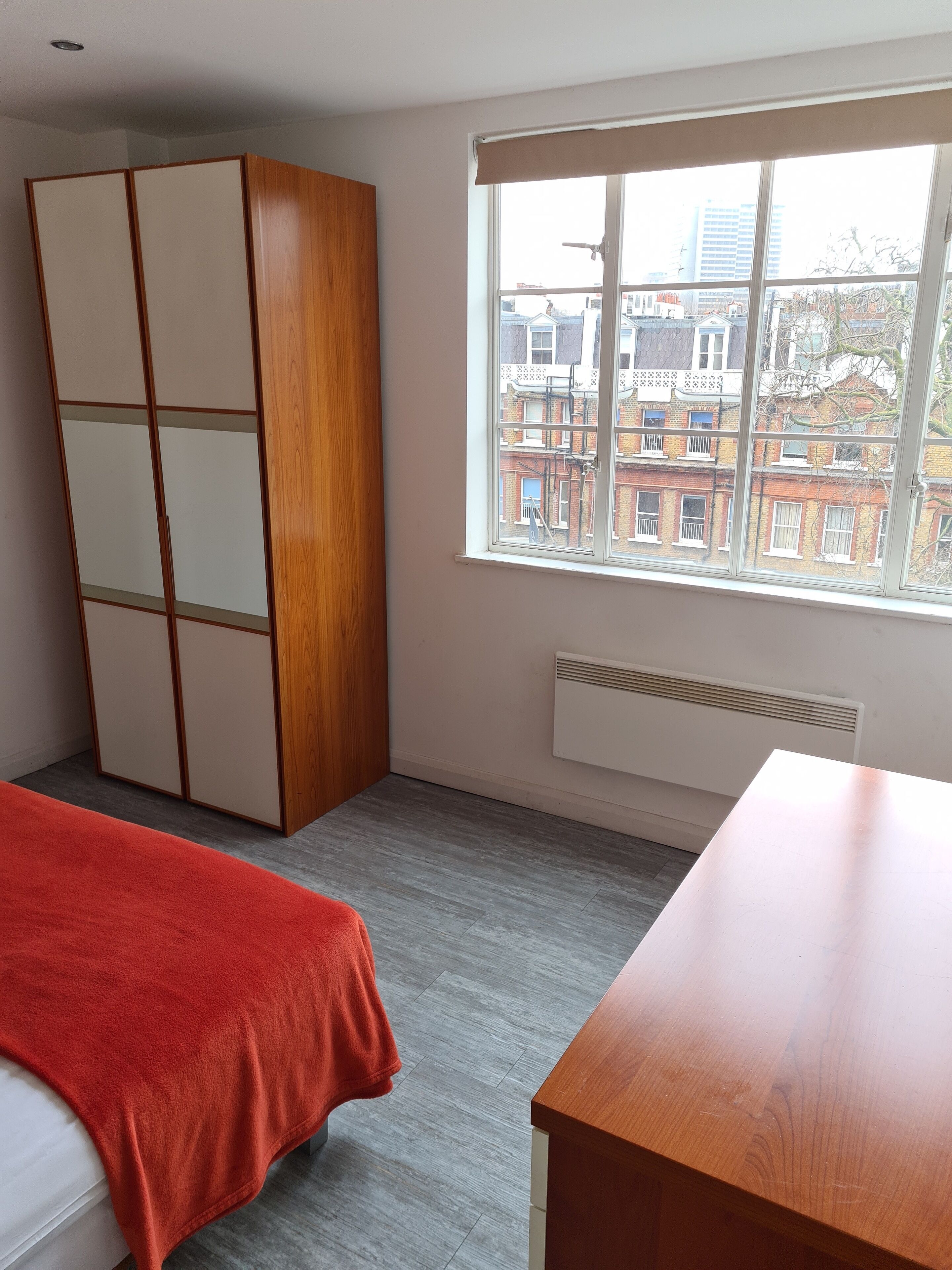 A bedroom with a bed, a wooden wardrobe, a window with a city view, and a radiator.
