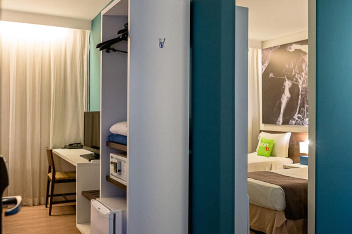 Une chambre d’hôtel de taille réduite, comprenant un lit simple, un bureau, une armoire et un miroir qui reflète la pièce.