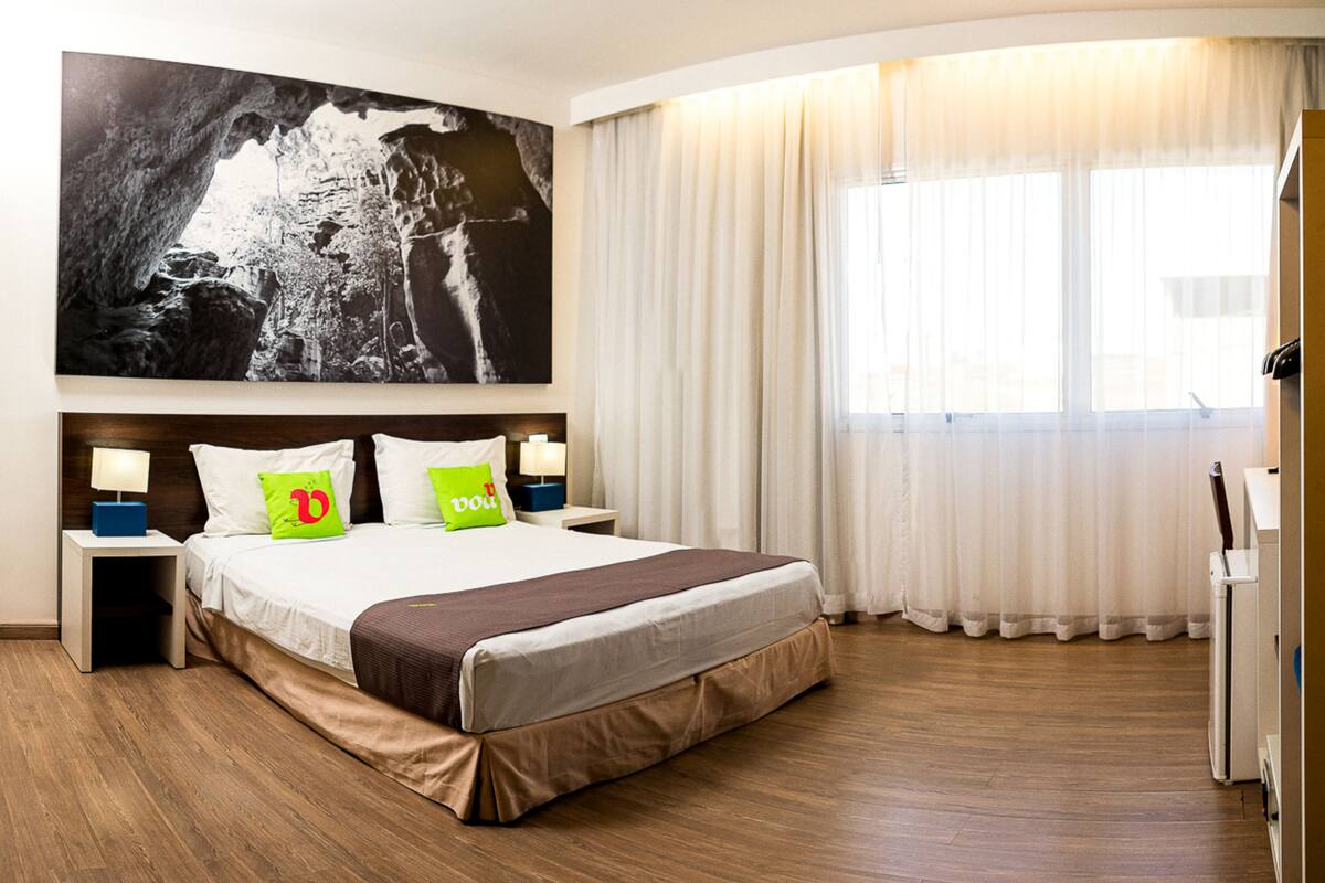 Ein Hotelzimmer mit einem großen Bett, zwei Nachttischen mit Lampen, einem großen Gemälde an der Wand und einem Fenster mit Vorhängen.
