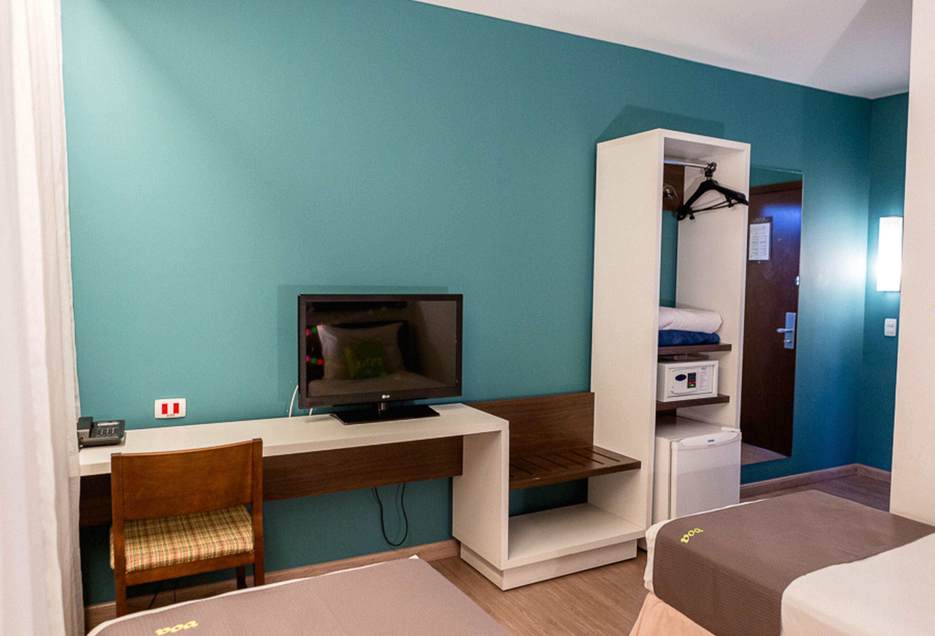 Ein Hotelzimmer mit einer türkisfarbenen Wand, einem hölzernen Schreibtisch mit einem Computer, einem Stuhl, einem Bett und einem Kleiderschrank.