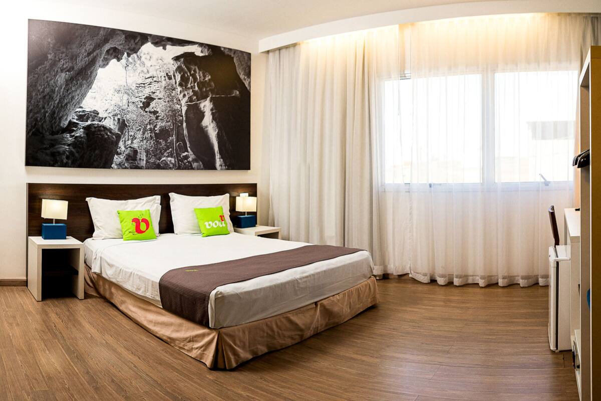 Ein Hotelzimmer mit einem großen Bett, einem Nachttisch mit Lampe, einem an der Wand montierten Fernseher und einem großen Fenster mit Vorhängen.
