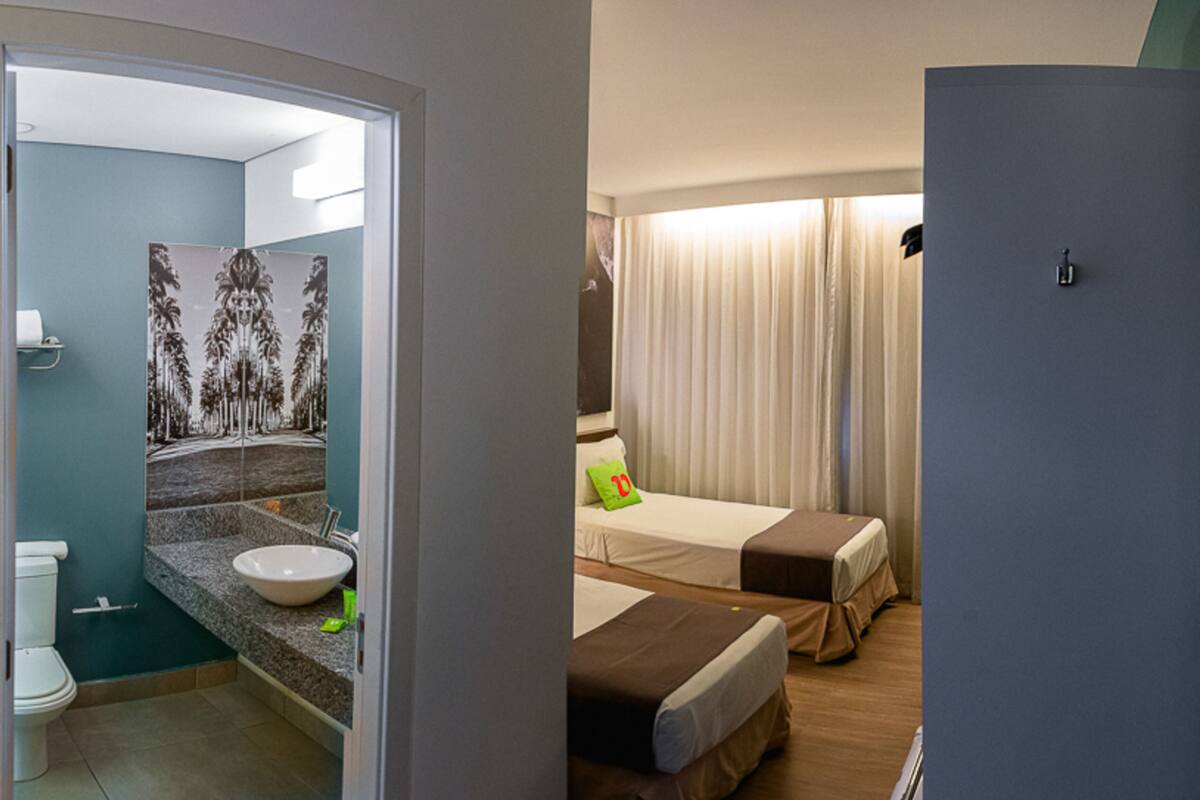 Ein Hotelzimmer mit Bad, das eine Toilette, ein Waschbecken und eine Wand mit Palmenmotiv zeigt. Das Zimmer verfügt über zwei Betten mit Kissen und ein Fenster mit Vorhängen.