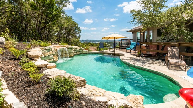 A backyard with a swimming pool, surrounded by rocks and plants, and a wooden deck with chairs and an umbrella. - Lake Travis Acreage with Hilltop Views  (Austin)