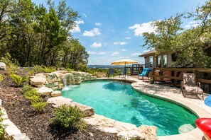 A backyard with a swimming pool, surrounded by rocks and plants, and a wooden deck with chairs and an umbrella. - Lake Travis Acreage with Hilltop Views (Austin)