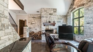 A spacious living room with a stone fireplace, leather seating, and a large window providing a view of the outdoors. - Lake Travis Acreage with Hilltop Views  (Austin)