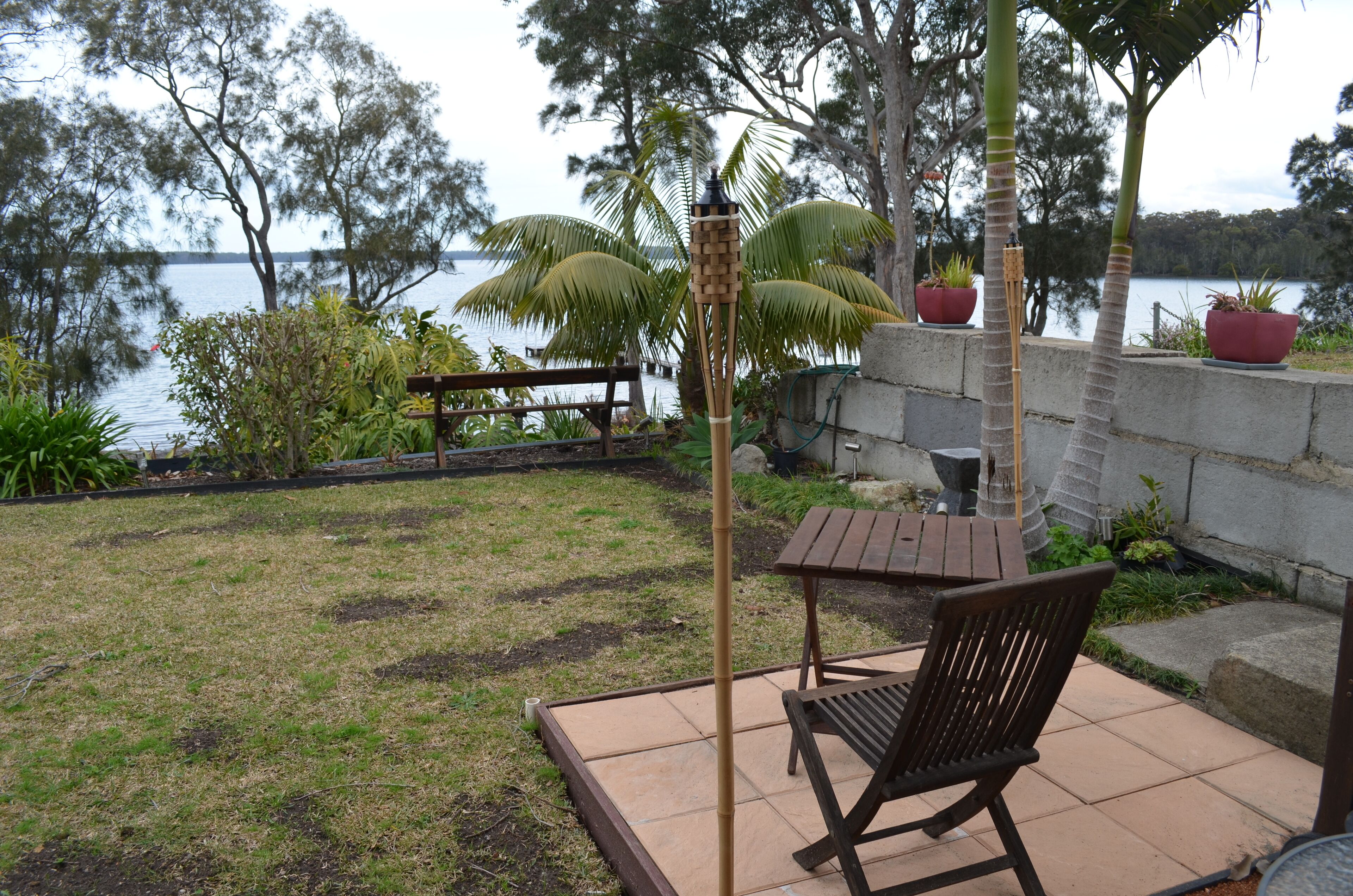 A patio with a wooden table and chairs, surrounded by potted plants and a view of a lake.