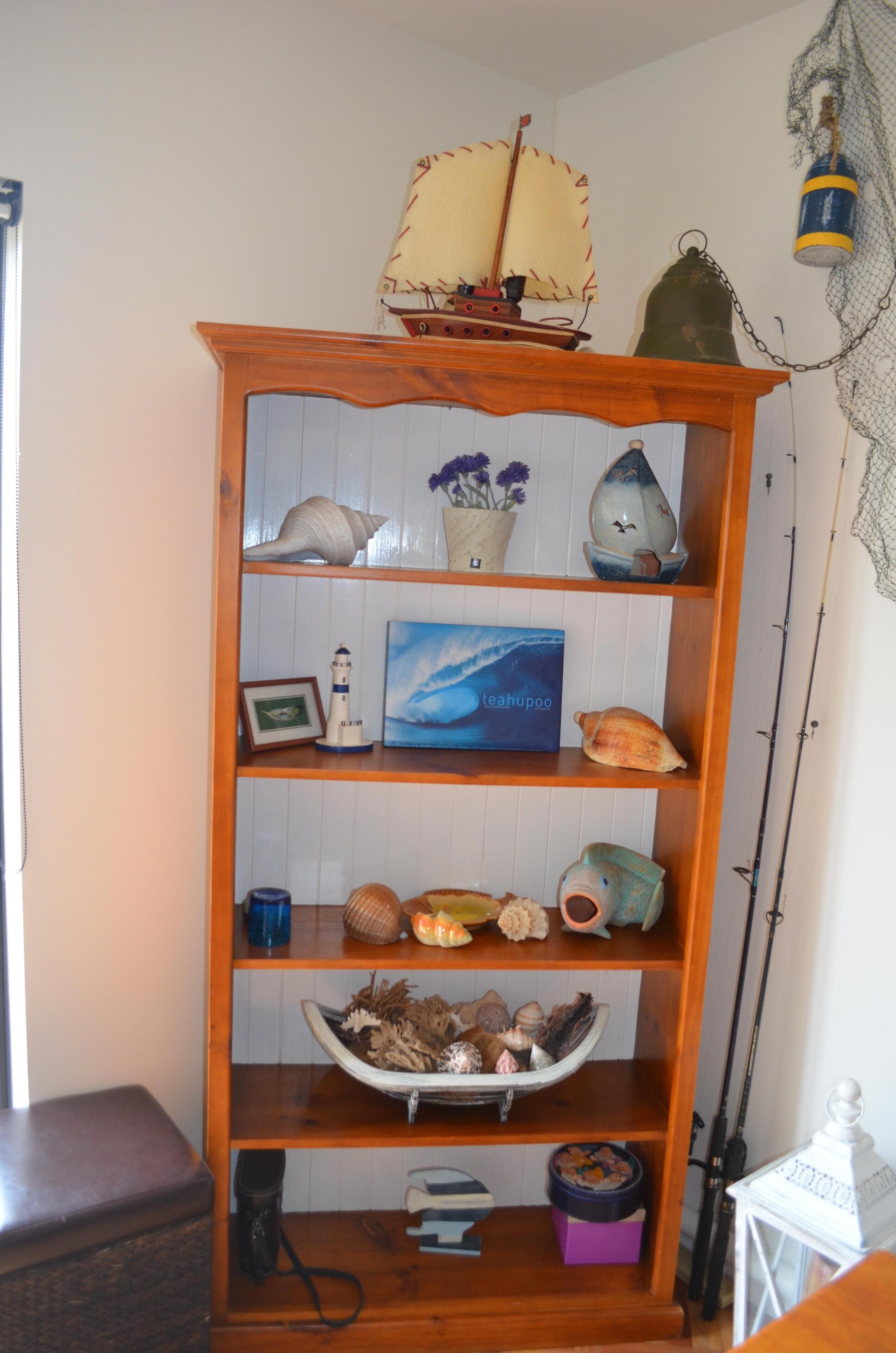 Uma estante de madeira com objetos decorativos, incluindo conchas do mar, uma réplica de navio e uma moldura com fotografia.