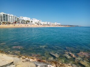 A beach with clear water, rocky shore, and white buildings in the background.