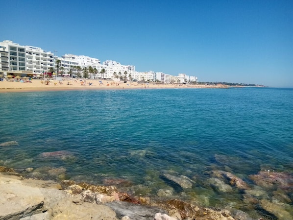 A beach with clear water, rocky shore, and white buildings in the background.