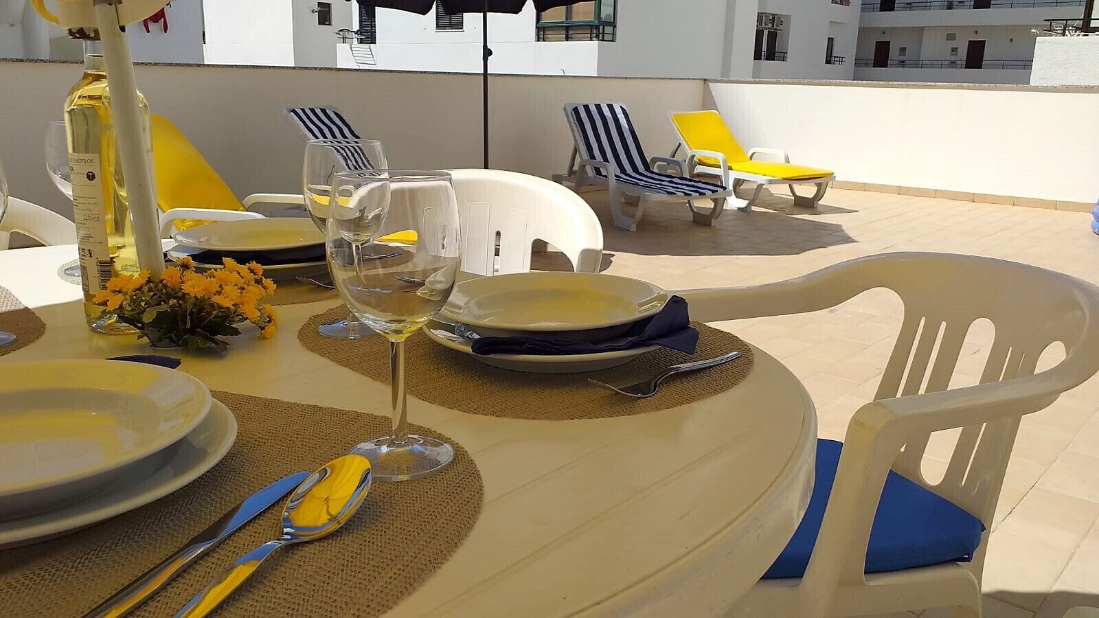 Een tafel voor twee, gedekt met borden, bestek en wijnglazen, omringd door lounge stoelen en een fles olijfolie.