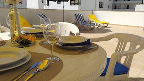 A table set for two with plates, cutlery, and wine glasses, surrounded by lounge chairs and a bottle of olive oil.