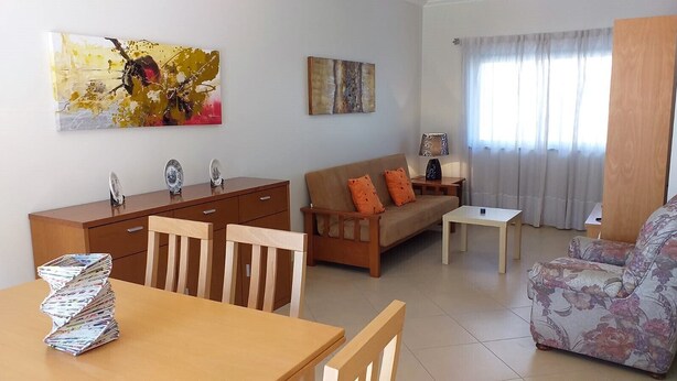 A living room with a sofa, a coffee table, a wooden dining table with magazines, a wooden cabinet, and two paintings on the wall. - Tahiti- Spacious, Private Parking, Terrace with Sunbeds, 300 meters Beach (Quarteira)