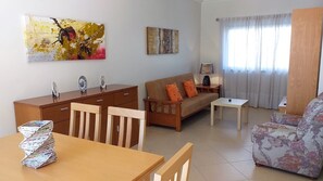 A living room with a sofa, a coffee table, a wooden dining table with magazines, a wooden cabinet, and two paintings on the wall.