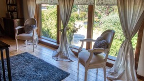 A room with two chairs facing a large window with curtains, a wooden floor, and a bookshelf.