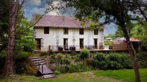 A two-story stone building with a balcony, surrounded by lush greenery and a wooden staircase leading to the entrance.