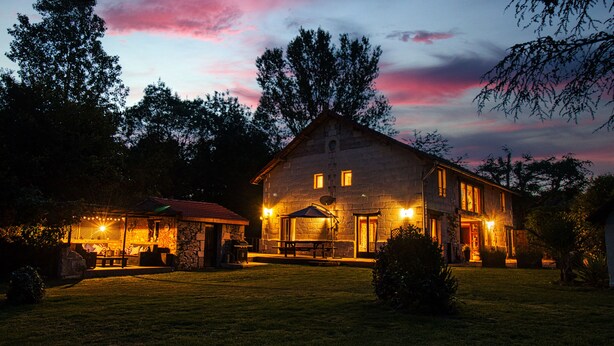 A stone building with a well-lit patio area, surrounded by trees and a grassy lawn.