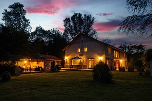 A stone building with a well-lit patio area, surrounded by trees and a grassy lawn.