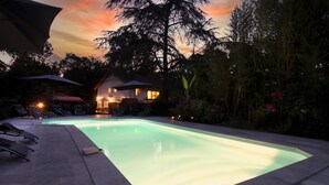 A swimming pool with a lighted edge, surrounded by lounge chairs and a house in the background.