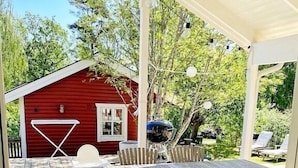 An outdoor patio with a wooden table and chairs, a red building in the background, and a pergola with string lights.