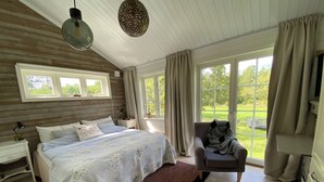 A bedroom with a large bed, a chair, a nightstand, a lamp, and a view of a garden through the windows. - 2 modern & cosy houses in Stockholm Archipelago 45 min. to Stockholm City.  (Norrtälje)