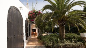 Un étroit sentier mène à un bâtiment blanc doté d’une grande porte en bois, d’un petit espace extérieur aménagé en coin salon et d’un palmier.