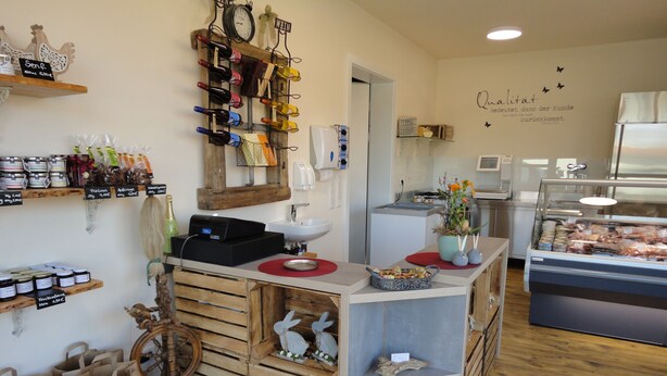 Interior de una cafetería con mostrador de madera, estantería para vinos y vitrina con productos alimenticios.