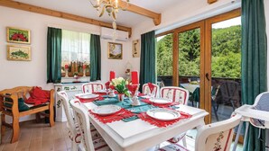 Ein Essbereich mit einem für sechs Personen gedeckten Tisch, Holzstühlen, einer Chandelüster und Blick ins Grüne durch große Fenster.