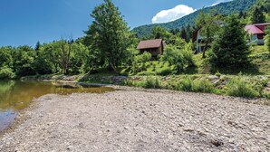 Blick auf einen Fluss mit einer Holzhütte und einem Haus, umgeben von üppigem Grün und Bergen.