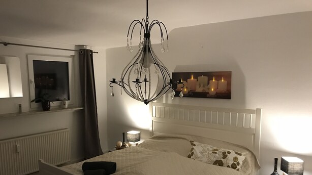 A bedroom with a white bed, a chandelier, a nightstand with a lamp, a mirror, and a wall-mounted picture. - Apartment Lichtblick near Centro  (Oberhausen)