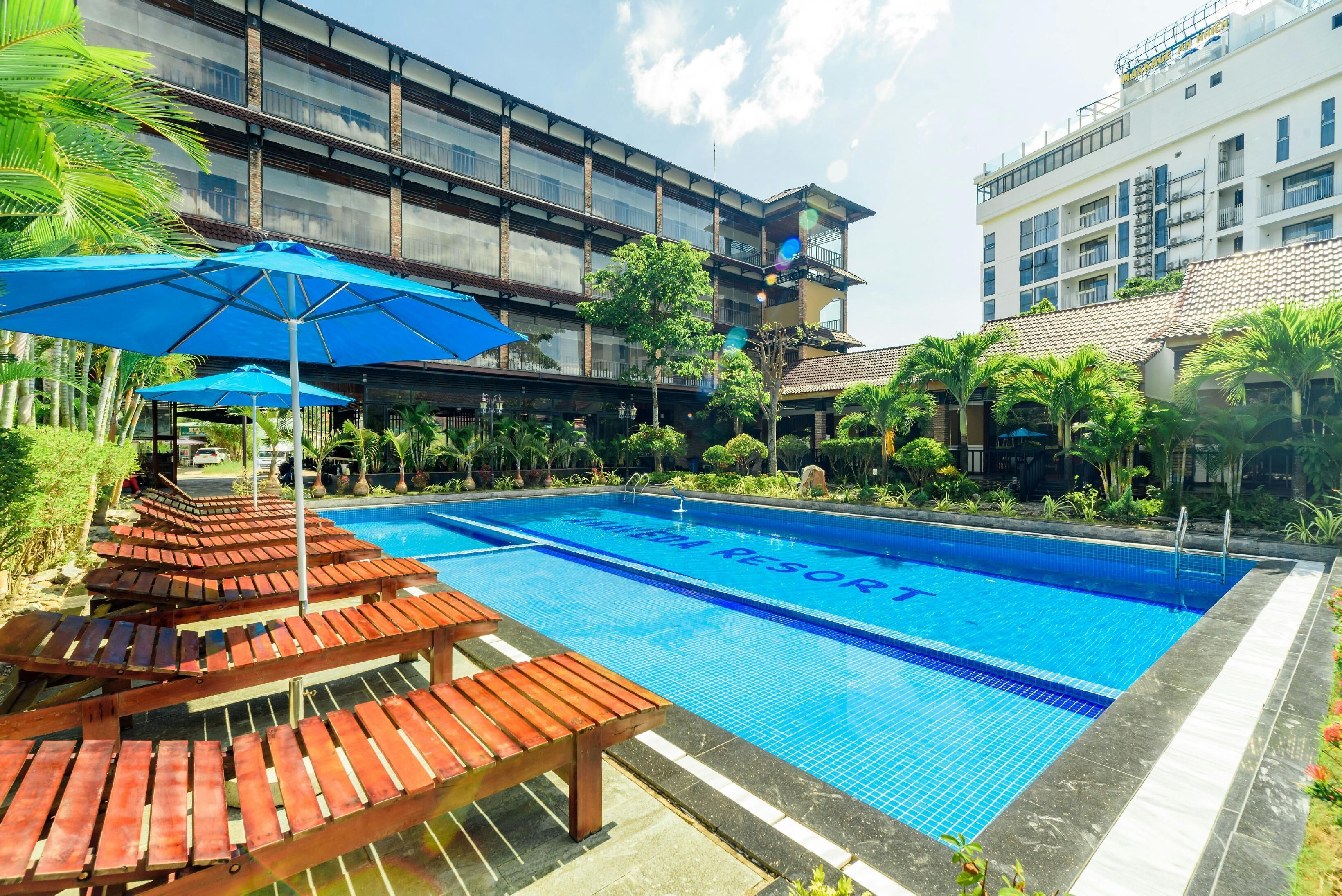 A swimming pool with wooden loungers and umbrellas, surrounded by greenery and buildings.
