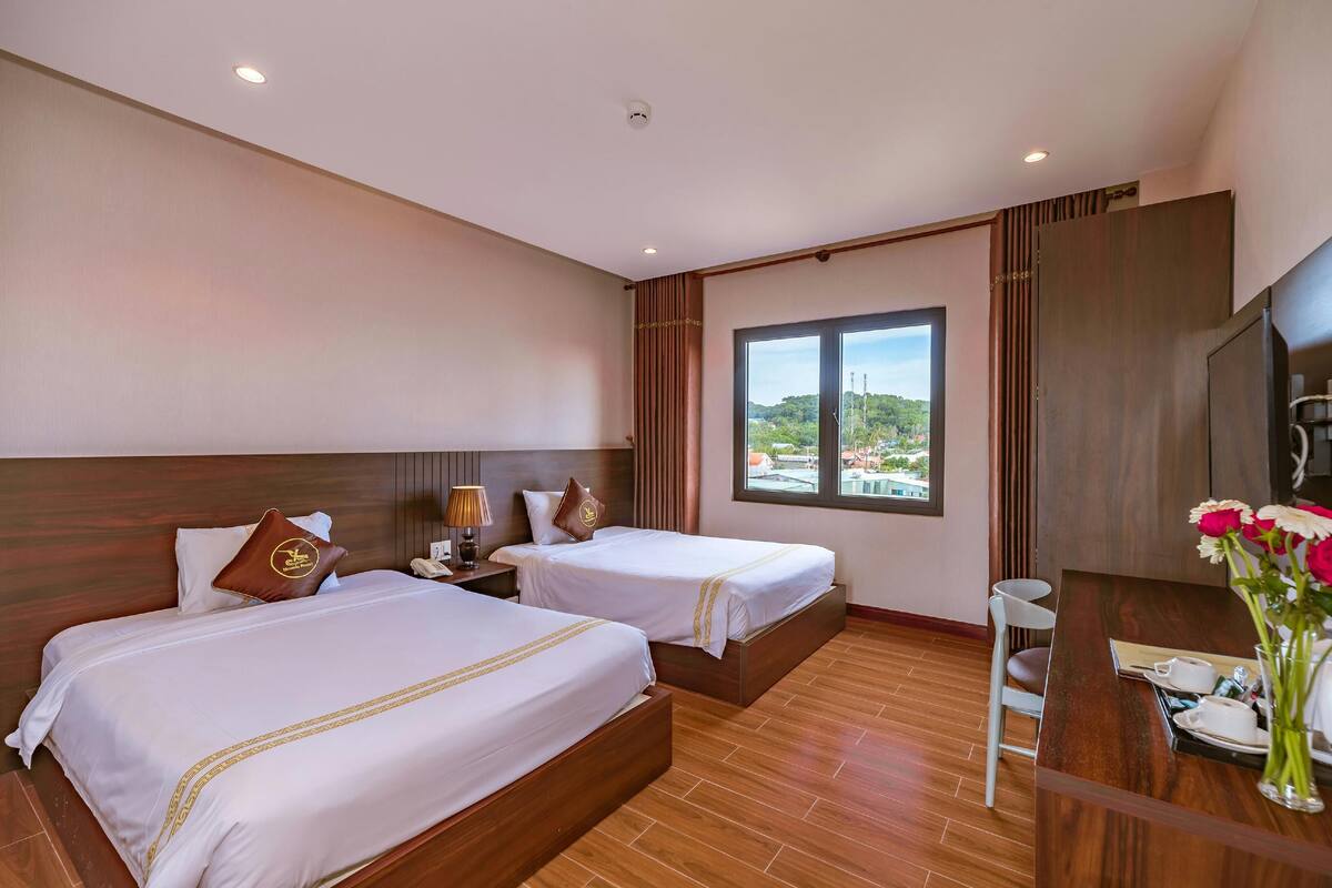 A hotel room with two beds, a wooden desk, a chair, a TV, and a window with a view.