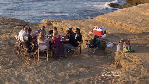 Een groep mensen dineert op een rotsachtig strand, met uitzicht op de oceaan.