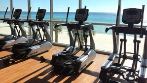 Une salle de sport équipée de machines de cardio et dotée de grandes fenêtres donnant sur une plage.