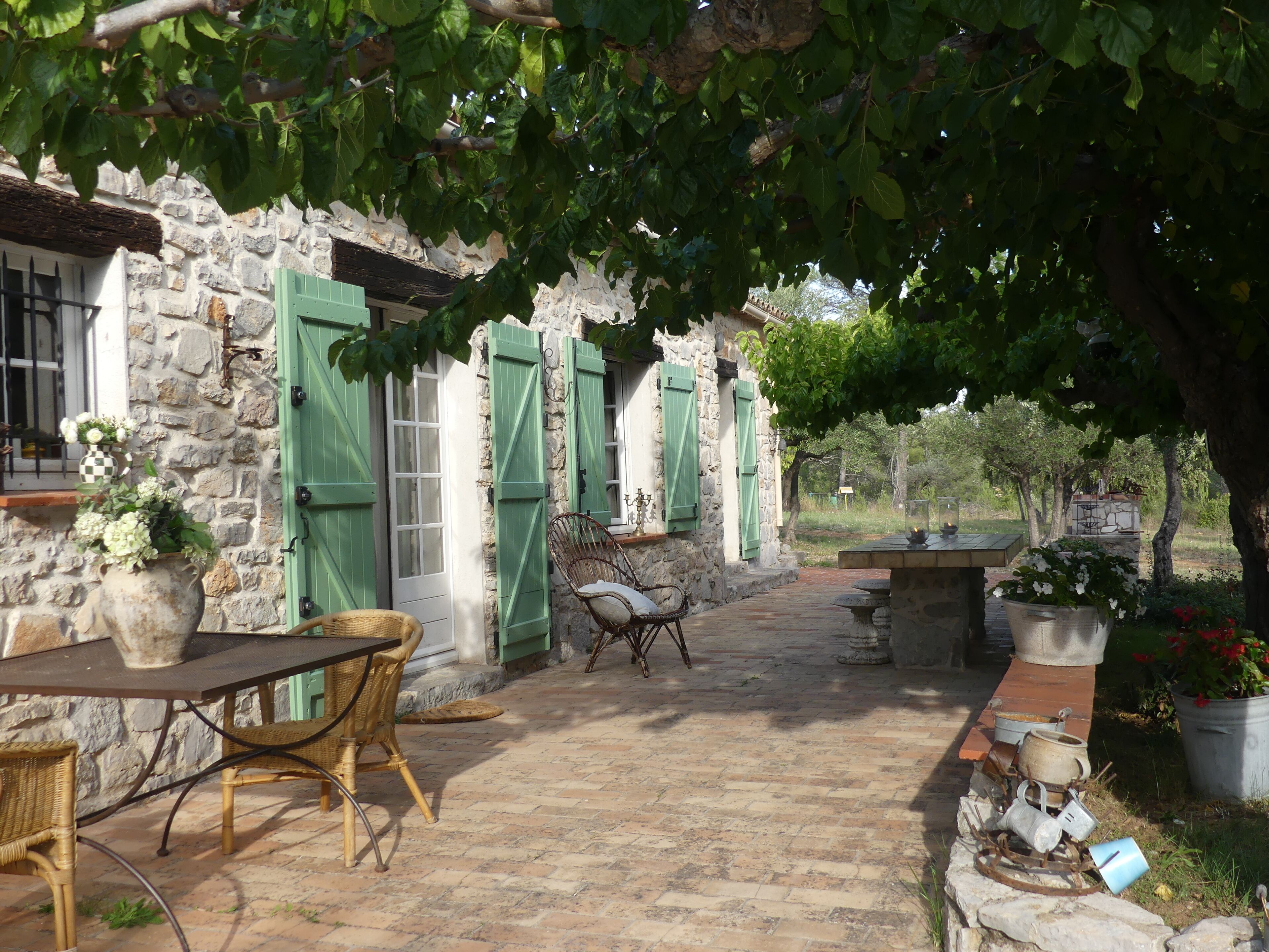 A stone building with green shutters, wicker furniture, and potted plants.